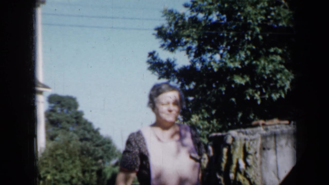 Woman standing outdoors in a sunny yard beside trees and a fence or low structure.