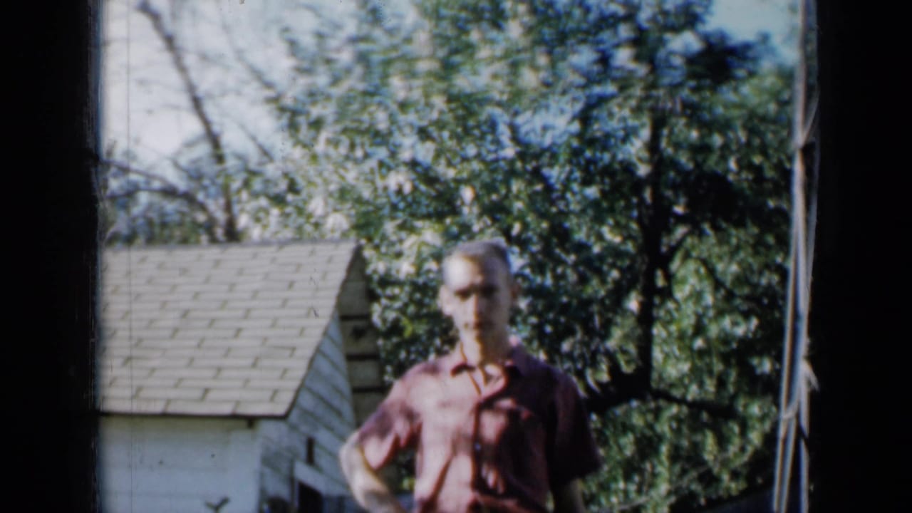 Man in a red shirt standing outdoors near a small shed and leafy trees.