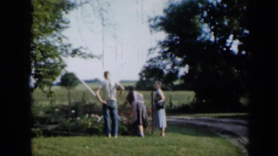 Three adults stand together near a grassy yard and garden edge, looking across open rural land in Minnesota.