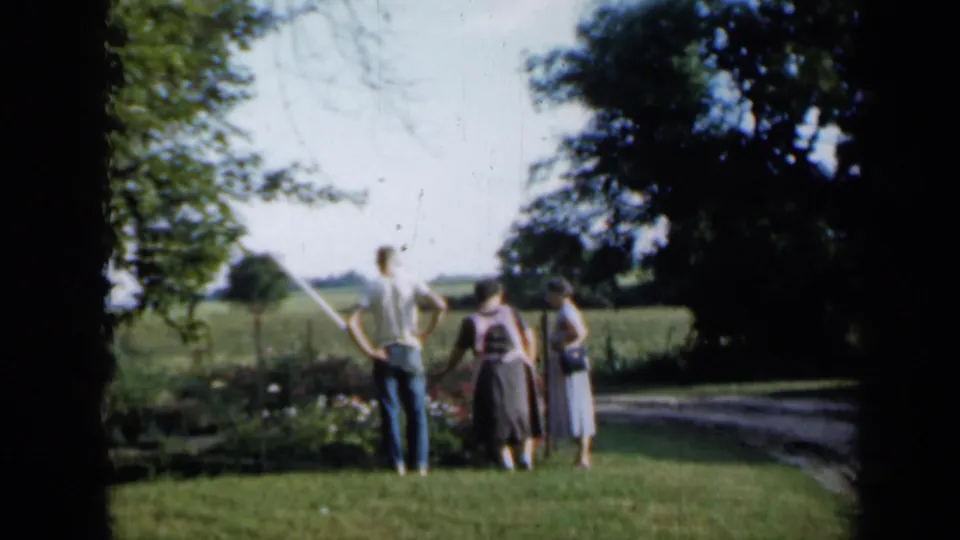 Two women and one man stand on a grassy verge near a curving rural road, with trees and fields in the background.