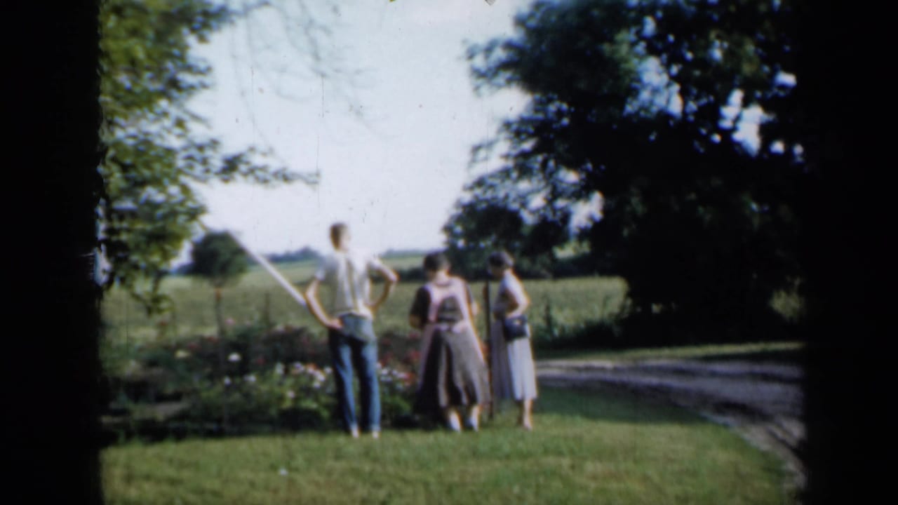 In metadata-supported 1958 Minnesota footage, a man and two women stand near a flower bed and fence with fields and trees behind them.