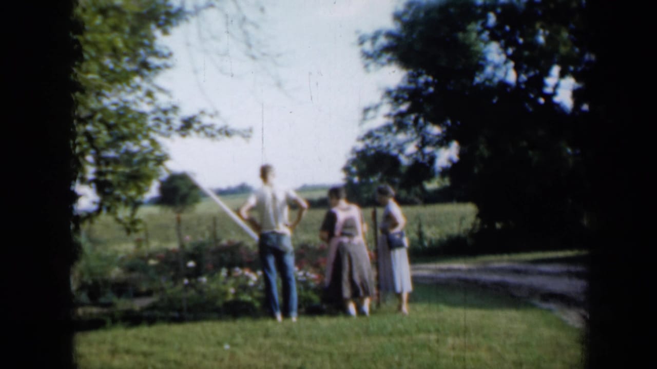 A man in a light shirt and two women in dresses stand beside a fenced flower bed in rural Minnesota, based on the clip metadata.