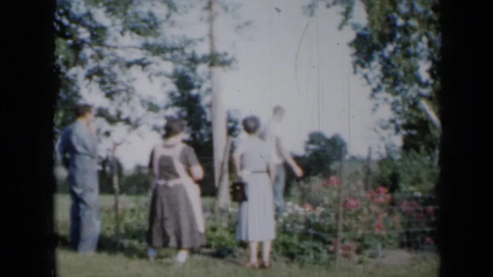 From behind, the adults face the garden and field, with a wire fence, flowers, and trees visible in this 1958 Minnesota home movie frame.