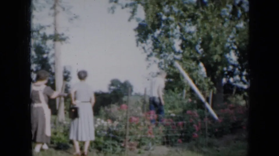 1958 Minnesota home-movie frame showing two women by a garden fence as a man tends flowers in a rural yard.