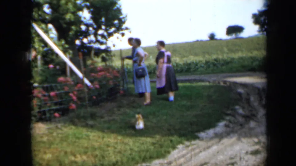 1958 Minnesota frame of women watching near a garden fence, with pink flowers, a small dog, and farmland beyond the yard.