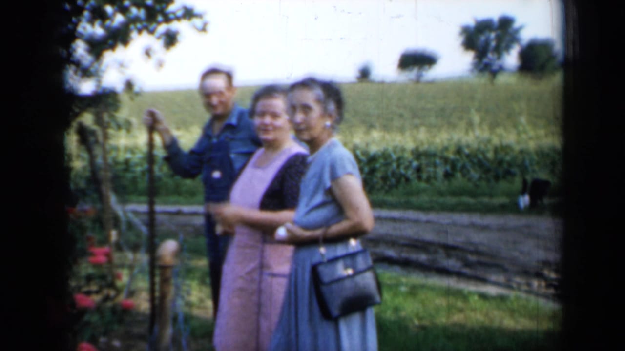 Three adults pose in a rural Minnesota yard, including two women in dresses and a man beside the garden fence.