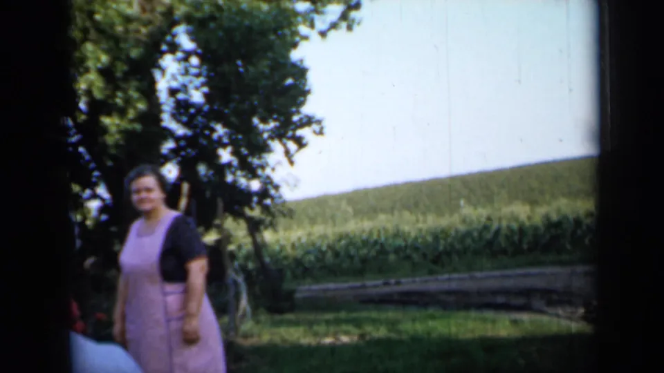 A woman in a pink apron stands in a sunny rural Minnesota yard with trees and a cultivated field behind her.
