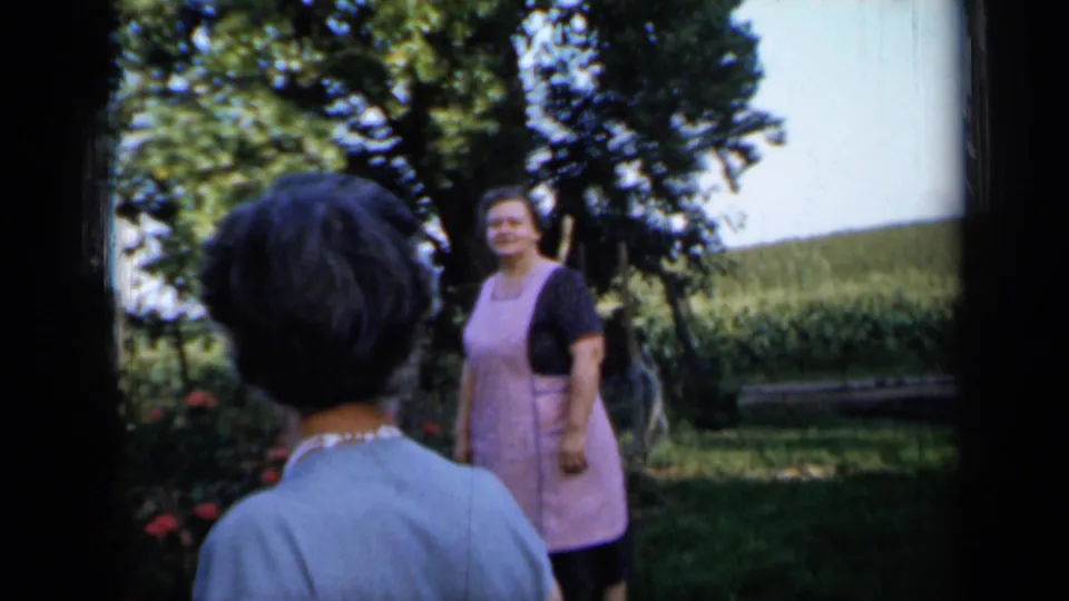 One woman is seen from behind while another woman in a pink apron poses near the garden in a 1958 rural Minnesota yard.