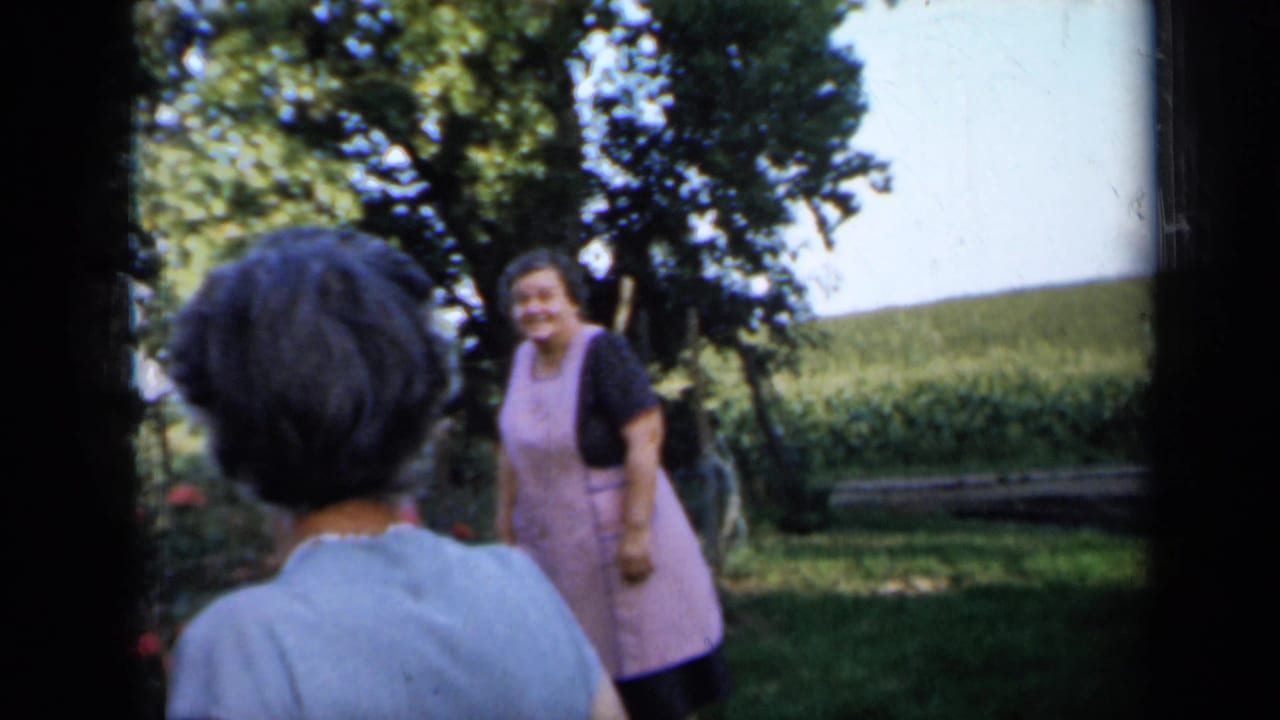 1958 Minnesota home-movie frame showing adults in a shaded backyard, including a smiling woman in a pink apron near a lawn and trees.