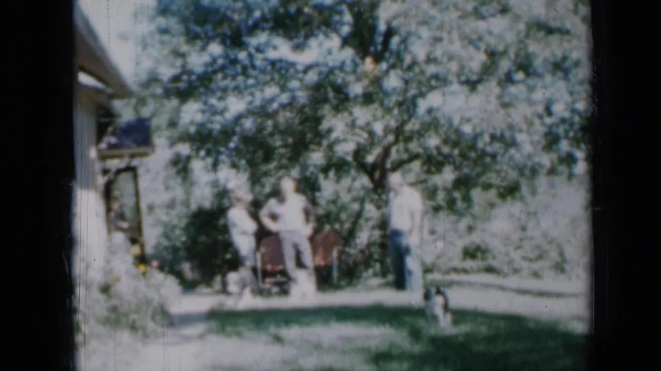 1958 Minnesota home-movie frame of adults gathered by a house and backyard doorway, partly shaded by a leafy tree.