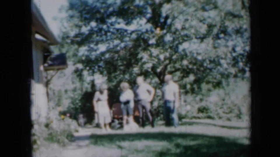 1958 Minnesota backyard gathering with adults arranged near a house, lawn, large tree, and garden plants in bright daylight.