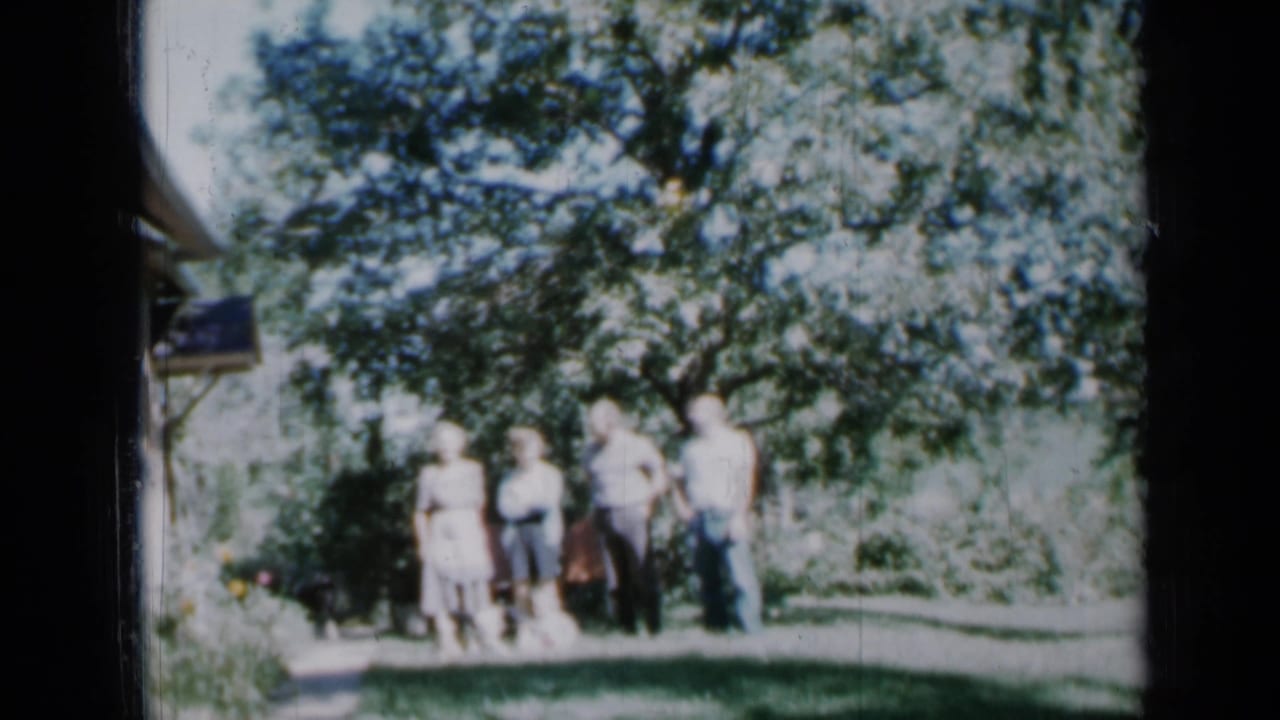 Minnesota 1958 frame of a small family group standing together outdoors in a backyard, partly shaded by a leafy tree.