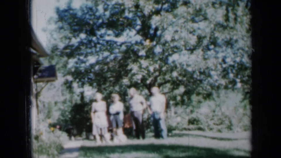 Minnesota 1958 frame of a small family group standing together outdoors in a backyard, partly shaded by a leafy tree.
