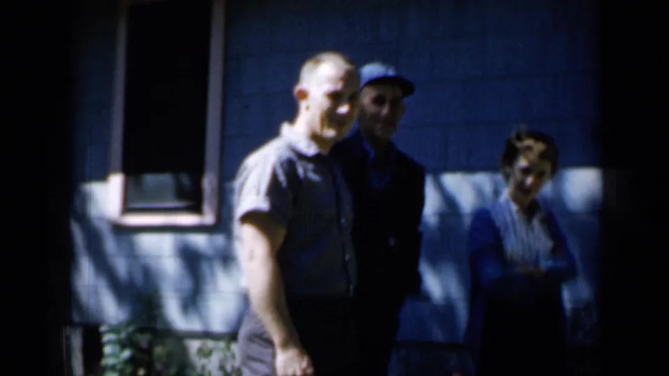 Closer Minnesota 1958 home-movie frame showing two men and a woman standing outside a blue-sided house.