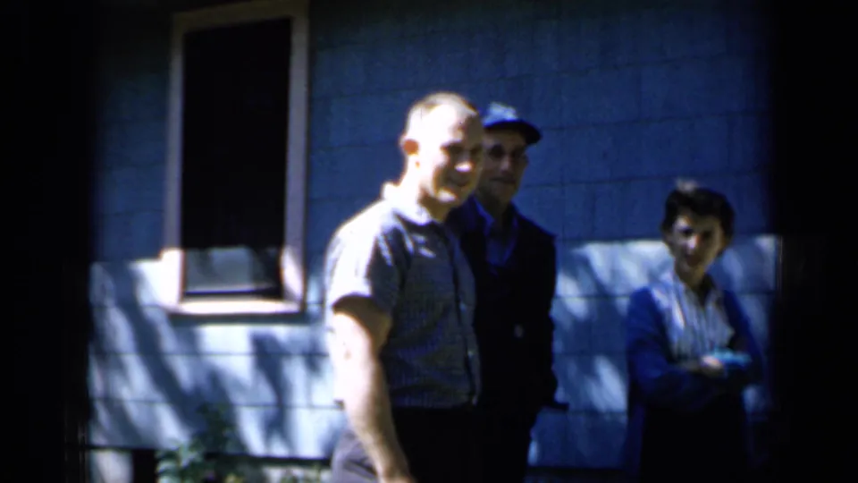 Minnesota 1958 frame showing three adults near a blue-sided house, with one man brightly lit in the foreground.