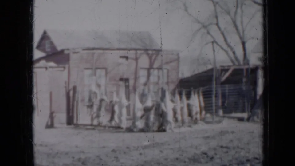 1958 Minnesota home-movie frame of adjacent small houses, a slanted-roof structure, fence posts, bare branches, and faint people.