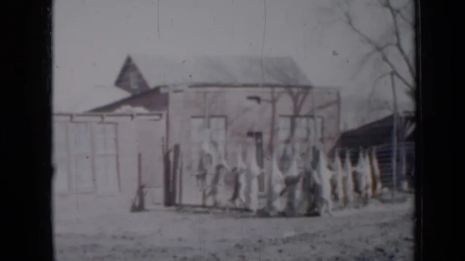 1958 Minnesota home-movie still showing a light-colored house, darker side structure, fence, bare trees, and faint people in the yard.
