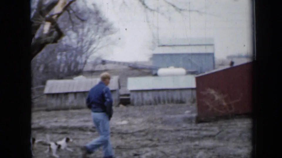 A man in a blue jacket walks across an open Minnesota farmyard as two dogs follow, with sheds, bare trees, and a red building edge visible.
