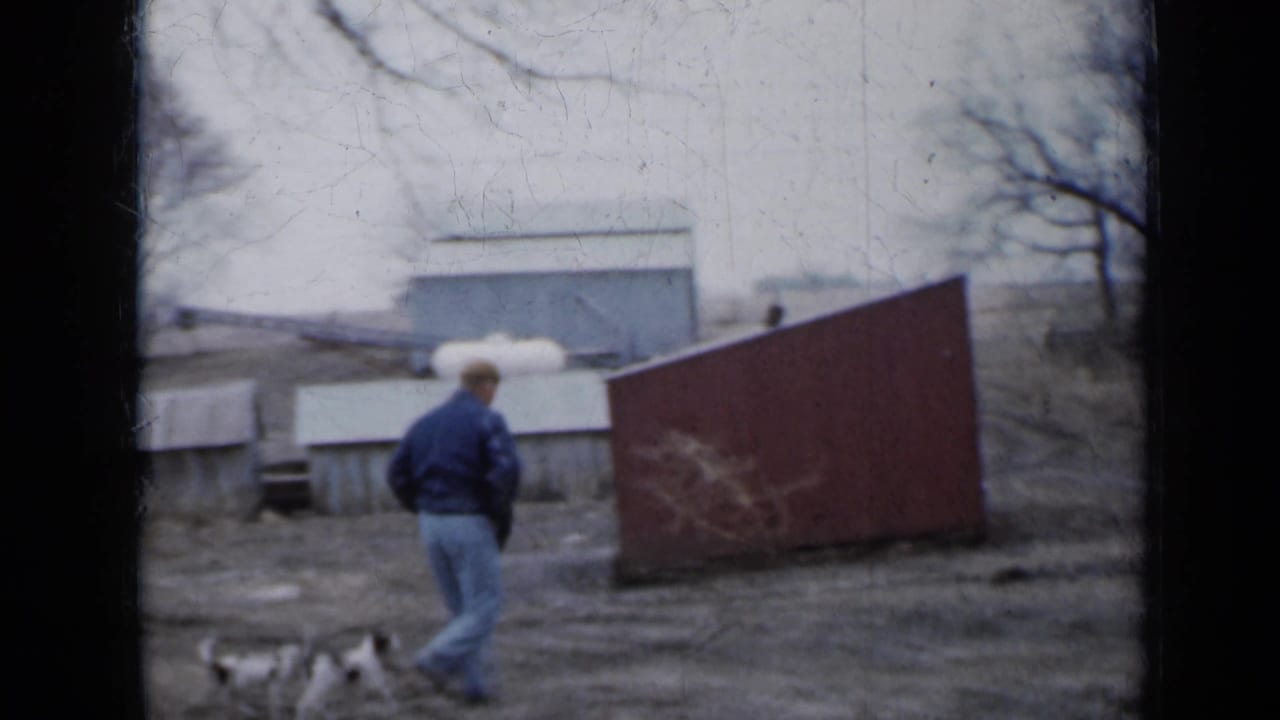 Man in a blue jacket walks across a muddy yard with small puppies nearby.