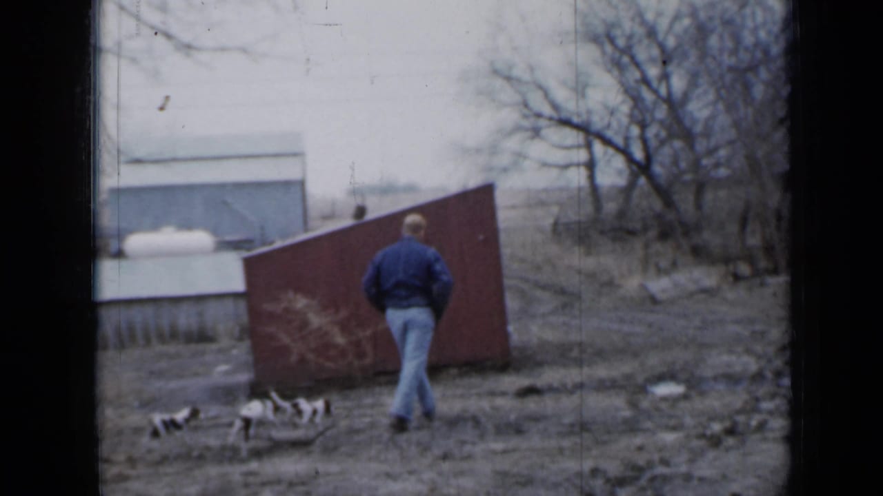 The man moves toward a red shed in a muddy Minnesota farmyard, with three black-and-white puppies clustered behind him.