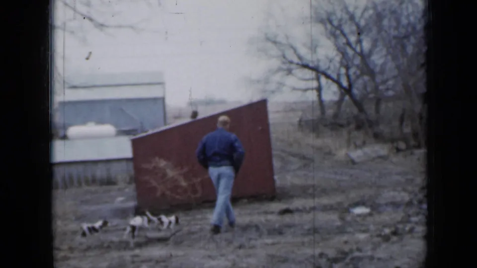 The man moves toward a red shed in a muddy Minnesota farmyard, with three black-and-white puppies clustered behind him.