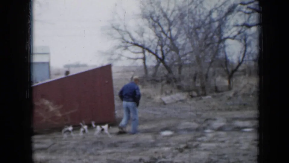 The man pauses near the black-and-white puppies beside the red shed, with bare trees and a muddy farmyard visible around them.
