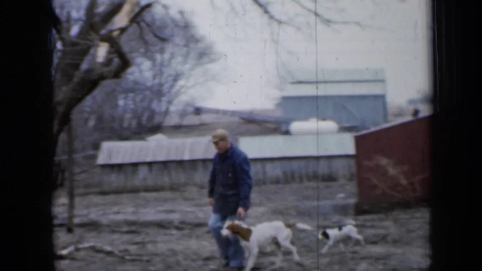 The man walks back through the muddy Minnesota yard beside a larger white-and-brown dog, with a small puppy, sheds, and propane tank behind him.