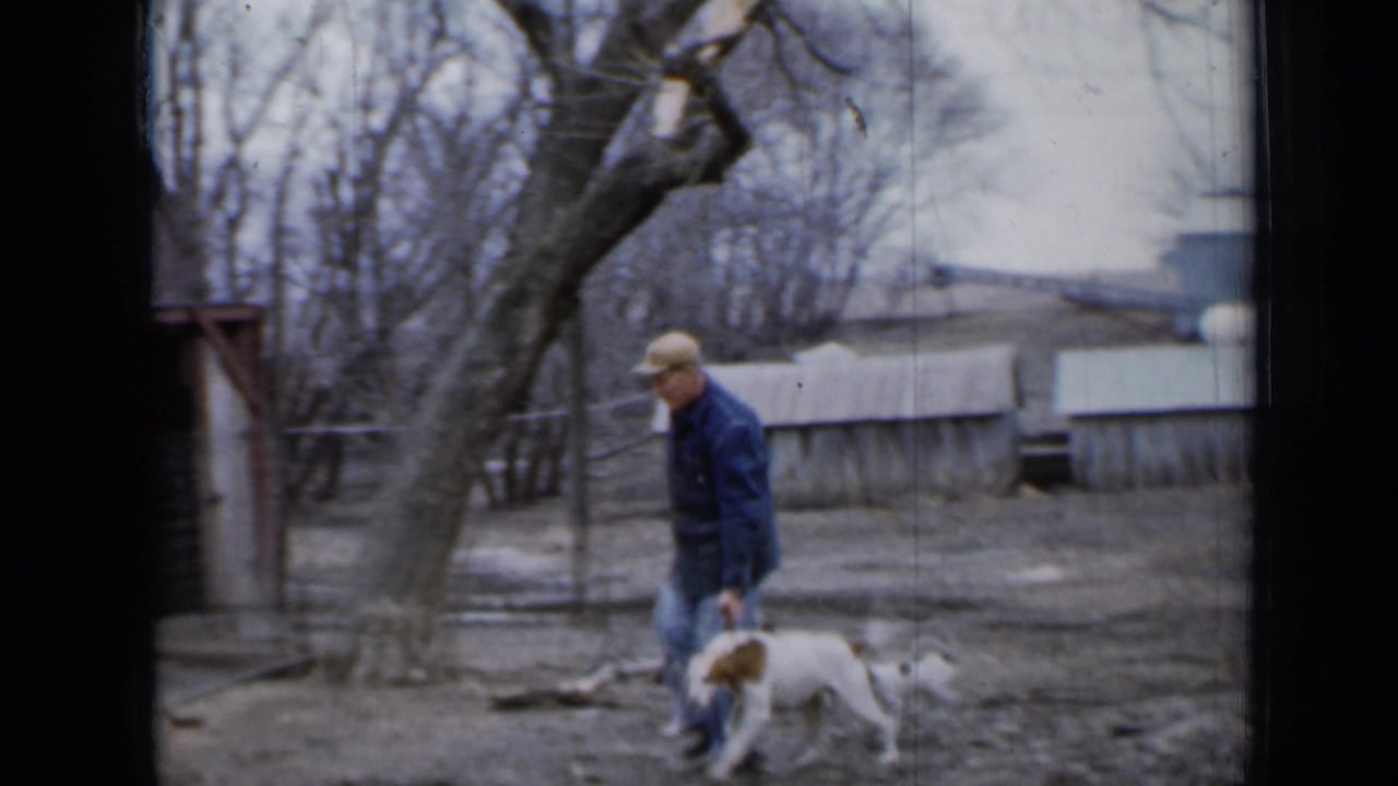 Man in cap walks a white and brown dog across a muddy farmyard.