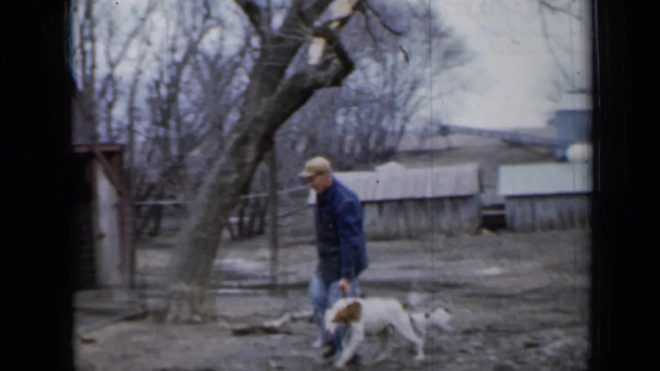 In 1958 Minnesota home-movie footage, a man in a blue jacket walks with a white and brown dog past sheds and bare trees.