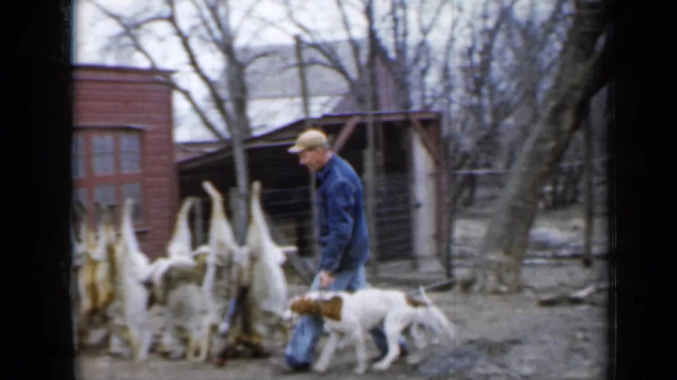 The man and dog pass a rack of skinned animals in a Minnesota farmyard, with a red outbuilding and bare trees behind them.