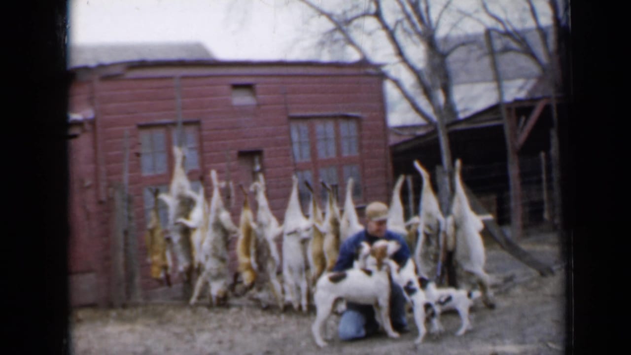 Hunter kneeling with dogs in front of hanging game and a red outbuilding.