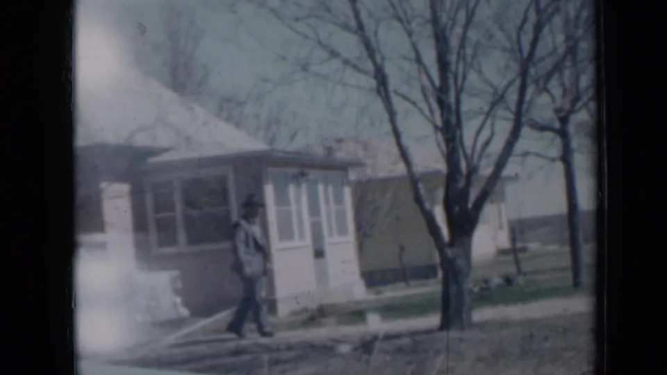 1958 Minnesota home-movie frame showing a person walking near a small house, yard, and bare tree.
