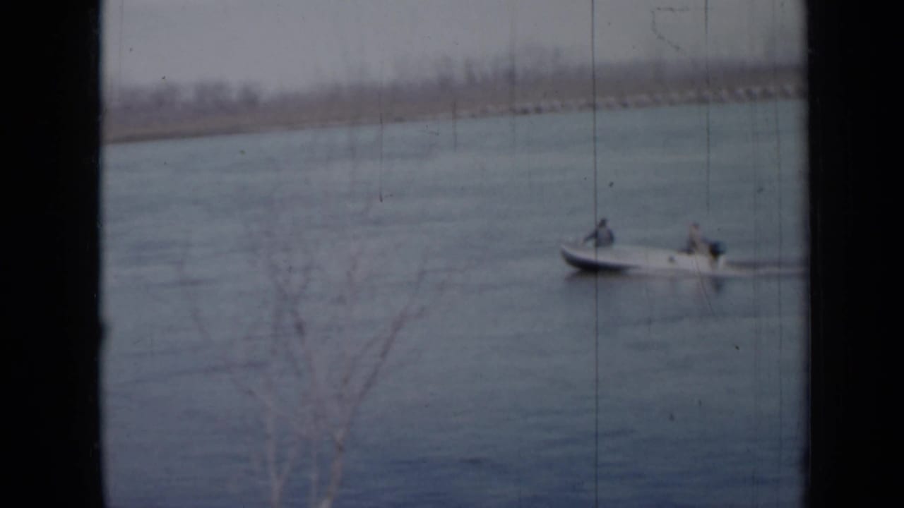 1958 Minnesota 8mm frame showing two people in a small motorboat crossing a calm lake beneath an overcast sky.