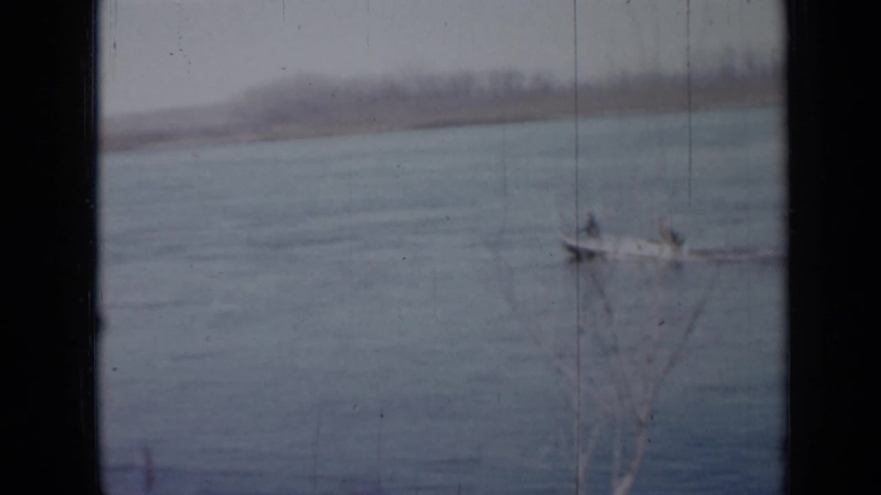 1958 Minnesota home-movie frame of a small boat with two distant occupants on a muted blue-gray lake.
