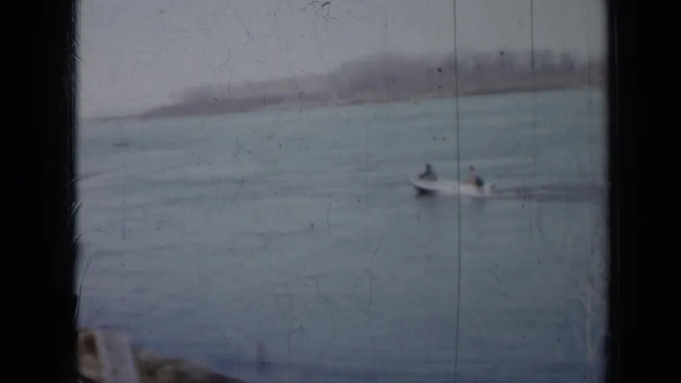 Two people in a small motorboat pass across a Minnesota lake in a soft, scratched 1958 8mm frame.