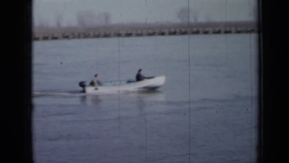 Small outboard boat with two occupants crosses calm Minnesota lake water in a 1958 8mm home movie.