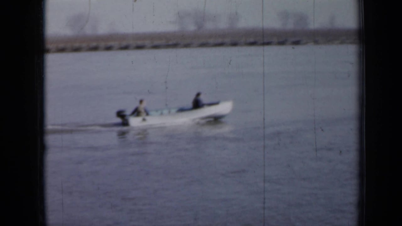 Small motorboat carrying two men moves across calm water near a distant shoreline.