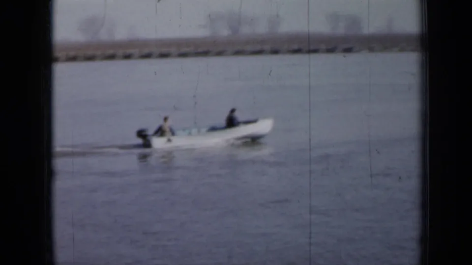 In this 1958 Minnesota home-movie frame, two men ride in a small outboard motorboat across calm river water.