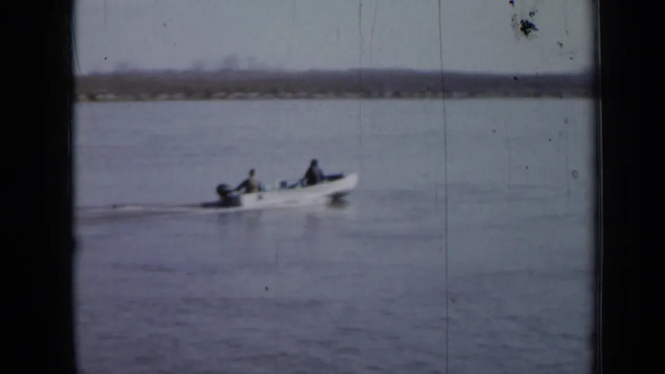 A small motorboat with two men aboard passes over calm water, with a low shoreline in the background.