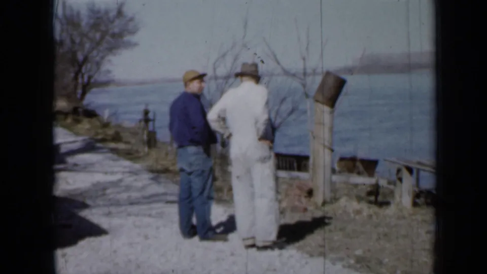 Two men, one in a blue shirt and one in white overalls, stand talking beside a Minnesota riverbank path in 1958 footage.