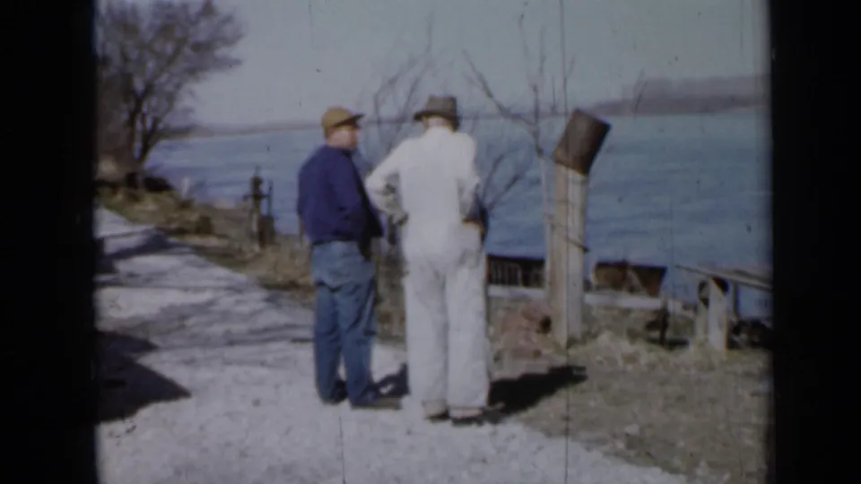 The shoreline view shows two men in workwear and hats standing by a path, with water and a distant bank beyond.