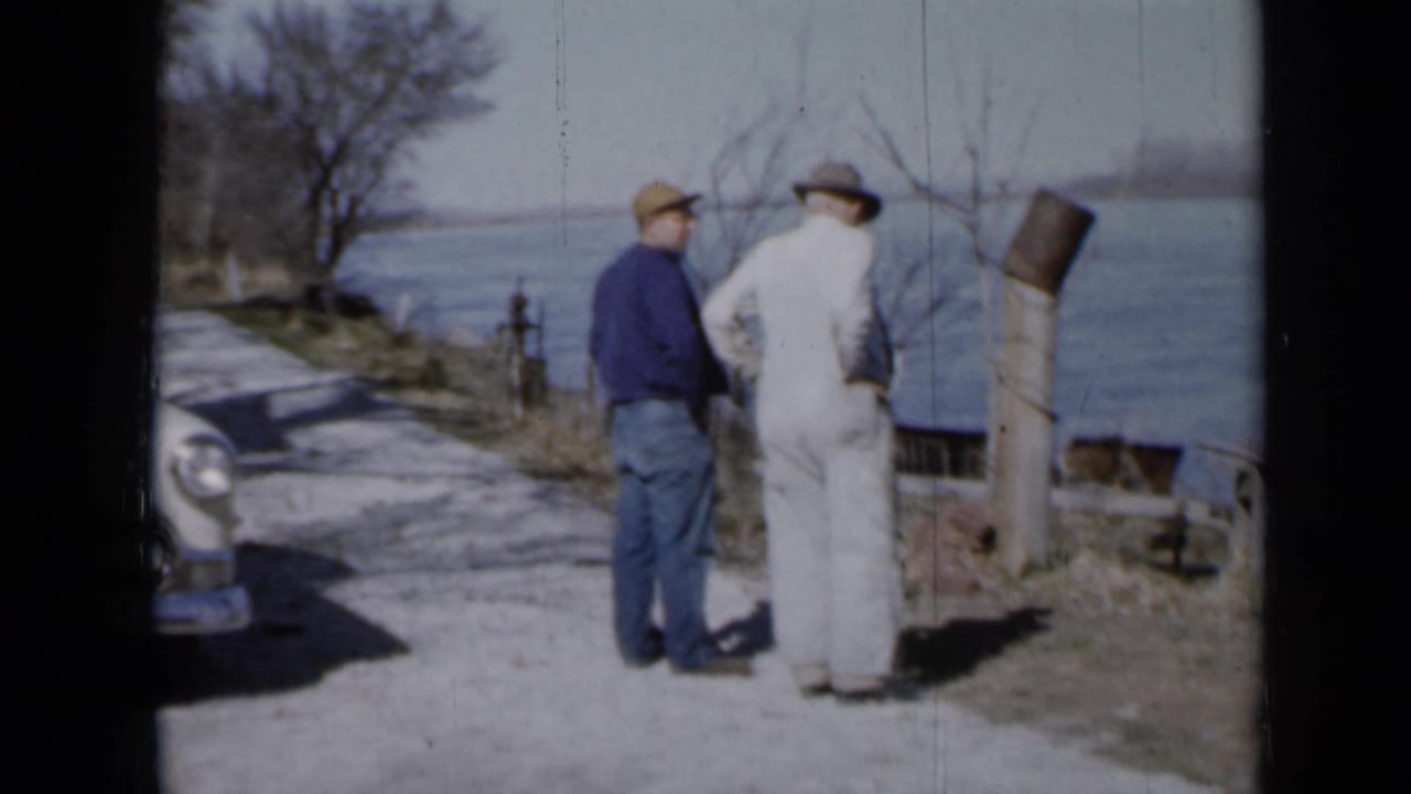 Two men in hats stand on a gravel path beside water with part of a car at left.
