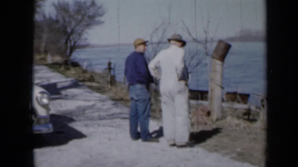 In stated 1958 Minnesota footage, two adult men stand on a riverside path near a partially visible vintage car.