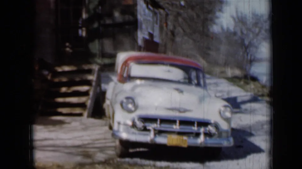 The front of a pale Chevrolet with chrome trim and red roof is framed near cabin steps in 1950s home-movie footage.