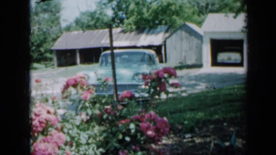 A 1958 Minnesota yard view shows pink flowers in front of a light blue car, lawn, trees, garage, and residence exterior.