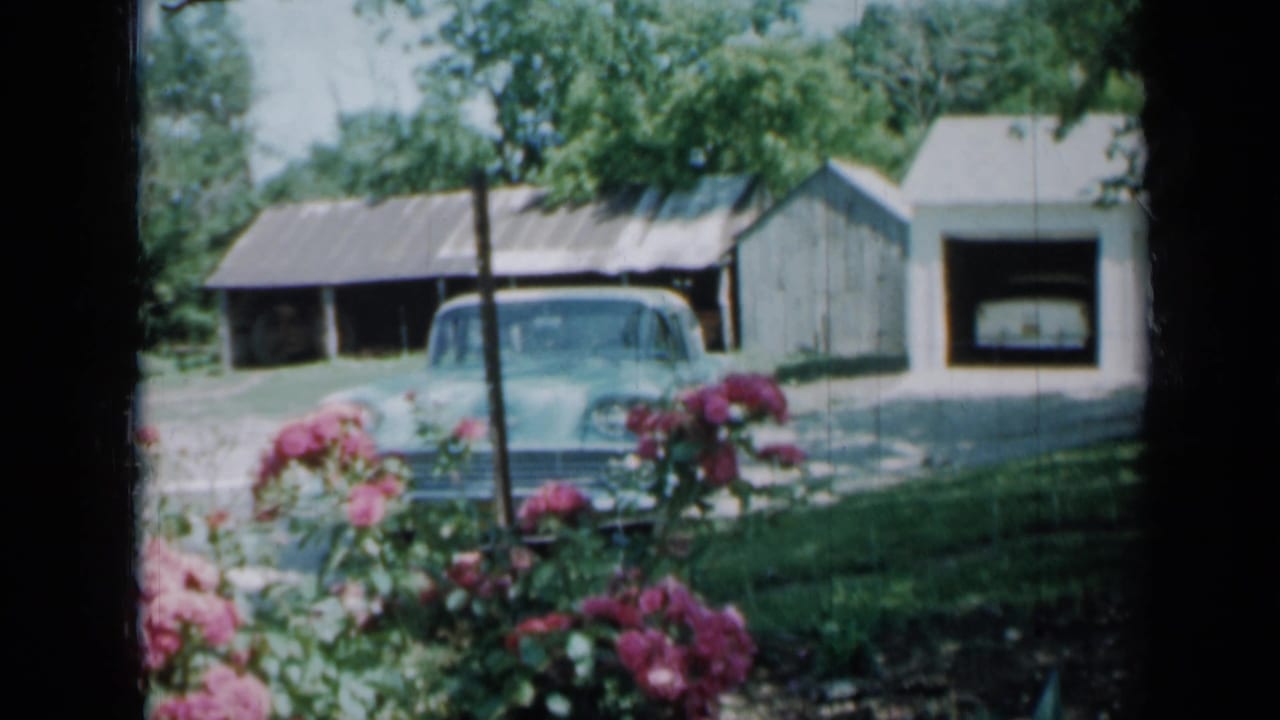 1958 Minnesota exterior with a parked light blue car, house, outbuildings, trees, and pink flowers in bright daylight.