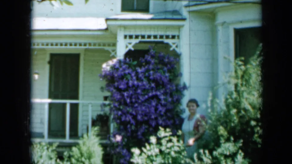 1958 Minnesota home-movie frame of a woman posed by a white porch and a large purple flowering vine.