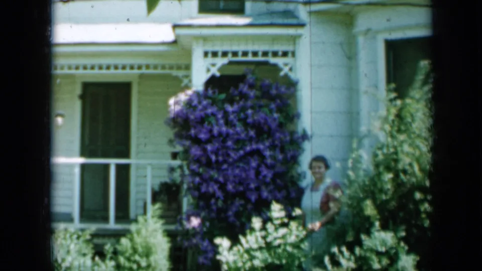 1958 Minnesota home-movie frame showing the woman near porch flowers and a white house exterior.
