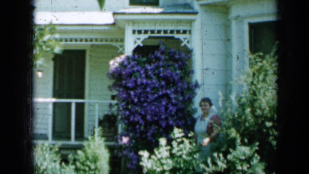 Woman standing in a garden beside a white porch and large purple flowering vine.