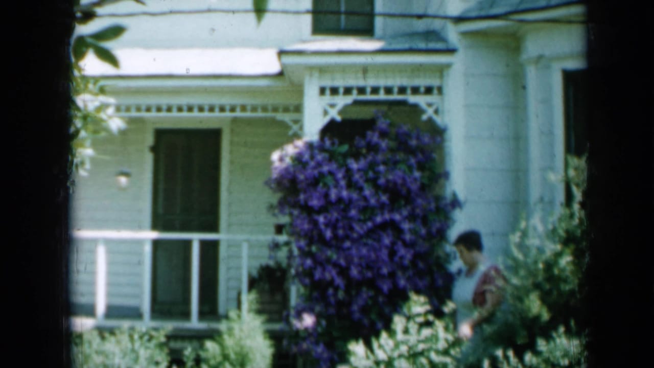 Woman partly visible near shrubs beside a white house porch and purple flowers.